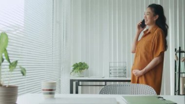 Asyalı hamile bir kadının ofiste tatildeyken cep telefonuyla iletişim kurduğu orta boy bir fotoğraf.