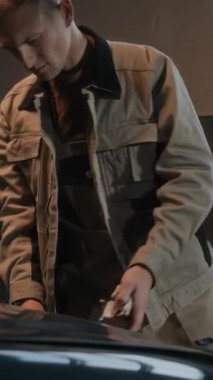 Garajda çalışırken üniformalı beyaz tamircinin araba kaputunu incelerken dikey görüntüsü.