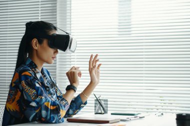 Genç Asyalı kadın ofisinde oturmuş, VR kulaklığını yeni bir proje için kullanıyor, boşluğu kopyalıyor.