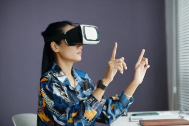 Orta boy Asyalı bir kadının şık bluz giydiği ve sanal uygulama kullanarak ofiste çalışan VR kulaklık taktığı bir fotoğraf.