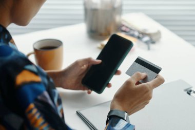 Online alışveriş yapan, akıllı telefon ve banka kartlarındaki uygulamaları kullanan, iş yerinde mola veren, tanınmayan genç bir kadının omuz üstü fotoğrafı.