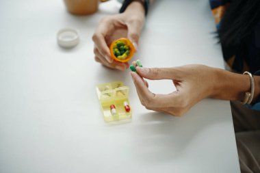 Yukarıdan bakıldığında tanınmayan kadın ellerinin masasında otururken ilaç düzenleyicisine vitamin kapsülleri eklediği görülüyor.