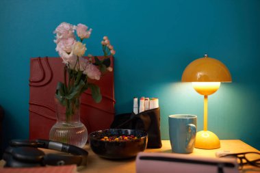 Vazoda çiçekler, kasede şekerler, lamba, keçeli kalemler, kupalar ve mavi duvarlı arka planda kitap olan natürmort yaşam kompozisyonu