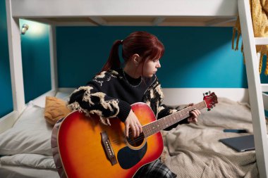 Z jenerasyonundaki kızın yurt odasında alt katta oturup gitar çalarken orta boy bir fotoğrafı.