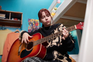 Z jenerasyonunun düşük açılı görüntüsü. Kafkasyalı kız yurttaki yatakta oturmuş gitar çalmayı öğreniyor.