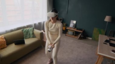 Beyaz aktif giysili kadın spor yaparken, evde gözlüklü ve hareket kontrolörlü artırılmış gerçeklik ekipmanı kullanırken yüksek açılı görüş.