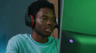 Çevrimiçi oyun oturumunda kapalı alanda bilgisayar başında mikrofon başında konuşan Afrikalı Amerikalı etnik kökenli mutlu bir adamın orta ölçekli yakın çekimi.