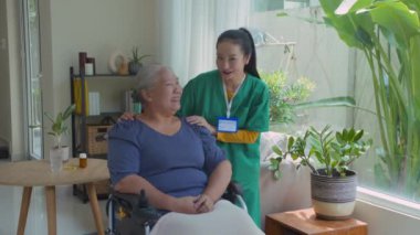 Tekerlekli sandalyede oturan gülümseyen Asyalı son sınıf öğrencisinin portresi. Pozitif genç bir bayan bakıcı, dairesinde elini omzuna koyuyor.