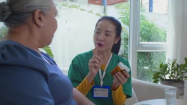 Tıbbi önlük giymiş Asyalı genç hemşire elinde iki şişe hap tutarken yaşlı kadın hastaya ilaç tedavisi uyguluyor.