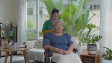 Tekerlekli sandalyedeki yaşlı kadının omuzlarına, ellerine ve bacaklarına dokunan Asyalı genç hemşire onu muayene ederken ve evde bakım ziyareti sırasında tablete not alırken