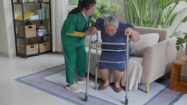 Yeşil önlük giymiş genç hemşire, kanepeden kalkıp yürüteç kullanma pratiği yapan Asyalı yaşlı bayana yardım ediyor.