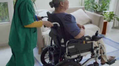 Yeşil önlük giymiş tanınmayan bir kadın bakıcı Asyalı yaşlı kadını tekerlekli sandalyede gezdiriyordu.