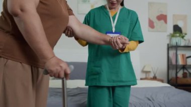 Asyalı genç kadın doktor felç üstüne destek için elini tutarken, tanınmayan yaşlı bir hastanın orta bölüm görüntüsü yavaşça bastonla yürümesi.
