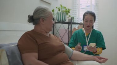 Kan basıncı monitörü kullanan ve evde bakım kontrolü sırasında Asyalı yaşlı bir hastayla sonuçları paylaşan arkadaş canlısı bir kadın bakıcı.