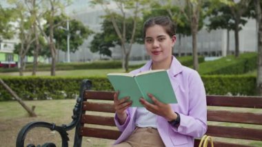 Sıradan giyimli neşeli çoklu etnik kadın portresi elinde kitapla bankta oturuyor ve parktaki kameraya gülümsüyor.