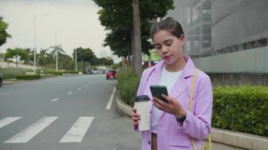 Lavanta ceketli Z jenerasyonunda elinde kahve, dışarıda taksi beklerken cep telefonunu kontrol eden çok kültürlü bir kadın.