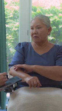 Tekerlekli sandalyedeki Asya 'lı yaşlı bayanın el eklem ağrısı hakkında konuşurken tablet üzerinde tanınmayan bir doktorun hastayı evinde ziyaret ederken yazması.