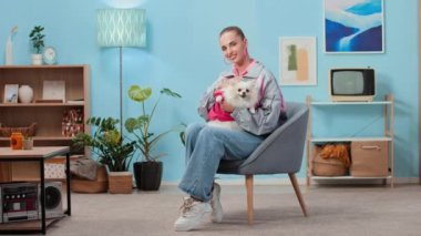 90 'lı yaşlarda gri ve pembe ceketli, kot pantolonlu ve spor ayakkabılı Pomeranya köpekli beyaz bir kadının uzun portresi. Koltukta oturmuş, retro oturma odasında kameraya gülümsüyor.