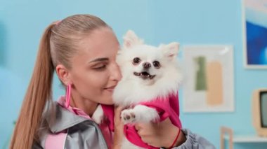 Beyaz tüylü köpeğini öpen pembe halka küpeler takan beyaz kadın milenyumun orta boy fotoğrafı.