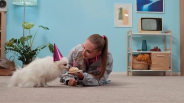 Y jenerasyonundaki beyaz bir kadının, retro oturma odasındaki kutlamalar sırasında doğum günü şapkalı sevimli tüylü köpeğe tatlı turta verirken geniş açılı görüntüsü.