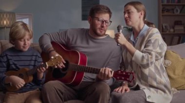 Üç kişilik beyaz bir ailenin mikrofona şarkı söylediği gitar çaldığı ve hafta sonu evde gitar tıngırdattığı orta boy bir çekim.