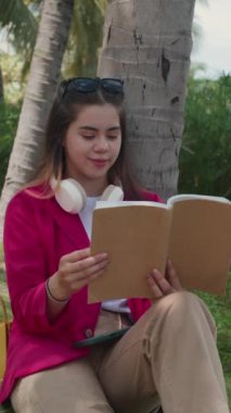 Şehir parkında şık giyinmiş, kitap okuyan bir kadının dikey görüntüsü.