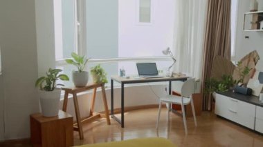 Güneş ışığıyla aydınlanan ev ofisinin iç kısımlarını pencere kenarındaki dizüstü bilgisayarla ve ev eşyalarıyla dolu rahat bir rafla çekmeyin.