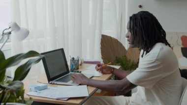 Genç siyahi erkek programcının laptop ekranına bakarken uygulama modelini incelerken ve ofisteki masada kağıt üzerinde notlar alırken orta uzunlukta bir kare.