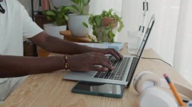 Akıllı telefon ve kablosuz kulaklıklarla masada otururken anonim Afrikalı Amerikalı iş adamının laptopta haber yaparken görüntüsü kesildi.