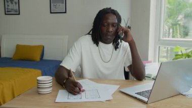 Afrika kökenli Amerikalı erkek grafik tasarımcısının akıllı telefondan ararken ve ev ofisinin bilgisayarını kullanırken uygulamayı kağıt üzerinde kontrol ederken orta boy görüntüsü.