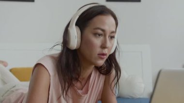 Kafkasyalı genç bir kadının kablosuz kulaklık takıp laptopta yazarken rahat bir yatakta dinlenirken çekilen görüntüsü.