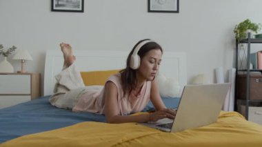 Kablosuz kulaklık takmış, laptopta çalışırken yatakta yatan Kafkasyalı genç bir kadın.