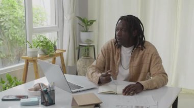 Genç Afrikalı Amerikalı bir adamın masa başında oturmuş, bilgisayarla dolu ev ofisinde çalışırken orta boy bir fotoğrafı. Not almak için defter açık.