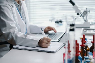 Bilim adamı araştırma yapıyor, verileri kaydediyor ve laboratuvar ortamında bilimsel çalışmalar için gelişmiş ekipmanlar kullanıyor. Çalışma alanında çeşitli laboratuvar aletleri ve kimyasallar görünür.