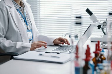 Laboratuvar önlüğü giyen kadın bilim adamı laptopta çalışıyor, etrafı laboratuvar ekipmanları ve kimyasallarla çevrili. Ekrandan bilimsel veri analizine odaklanan araştırmacı