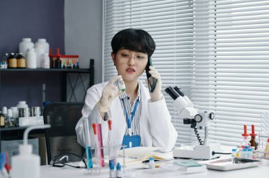 Laboratuvar önlüğü ve gözlük takan kadın bilim adamı, laboratuvar ortamında çeşitli bilimsel aletlerle telefonda konuşurken araştırma yapıyor.