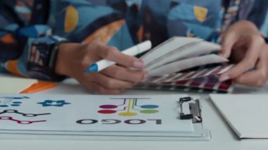 Yaratıcı çalışma alanındaki iyi organize edilmiş çalışma masasında canlı logo tasarımı için renk örnekleri ve çizimler inceleyen tanınmamış kadın tasarımcının ellerini yakın plan çekin.