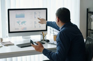 Modern ofiste akıllı telefonu elinde tutarken bilgisayar ekranında görüntülenen verileri gösteren bir adam. Grafikler ve haritalar analiz süreci ve çalışma alanı organizasyonu hakkında içgörü sağlıyor