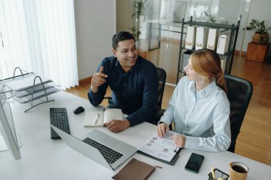 Erkek ve kadın sohbete dalıyor, belgeleri inceliyor ve ofis masasındaki bilgisayarlarda çalışıyor. Etrafı bitki ve ofis malzemeleriyle çevrili.