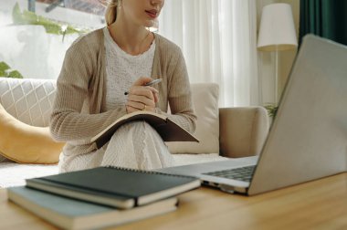 Not defterine yazarken ve elinde kalem tutarken koltukta oturan bir adam. Dizüstü bilgisayar ve diğer kitaplar masada ön plandayken defter ve kalem kucağa konur.