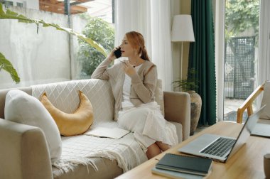 Modern oturma odasında kanepede oturan bir kadın, telefon konuşmaları ve bardakla meşgul. Geniş pencereler doğal ışık girmesini sağlıyor, rahat bir atmosfer yaratıyor.
