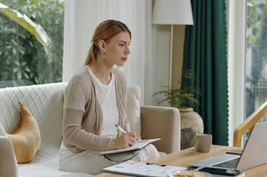 Tahta bir masada dizüstü bilgisayarla notlar üzerinde çalışırken rahat bir koltukta oturan bir kadın.