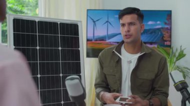 Genç Hintli erkek mühendisin mikrofonla konuşurken, rüzgar türbinleri üretimi hakkında podcast yaparken, yeşil enerji ofisinde gazetecilerin önünde otururken orta boy bir fotoğraf.