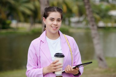 Portrait of young woman smiling at camera while drinking coffee and using digital tablet in the park clipart