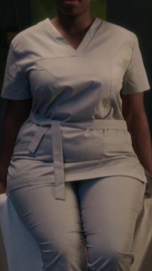 Profesyonel siyahi kadın spa terapistinin kamera önünde poz veren önlüklü portresi ve mesai saatleri boyunca masaj masasının kenarında oturan kolları dikey eğimli.