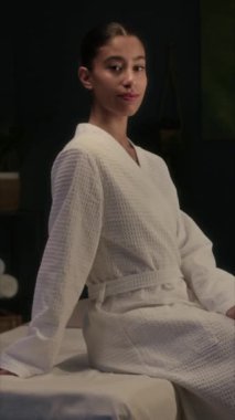 Beyaz bornozlu esmer kadının dikey portresi masaj masasında oturan kameraya gülümsüyor.