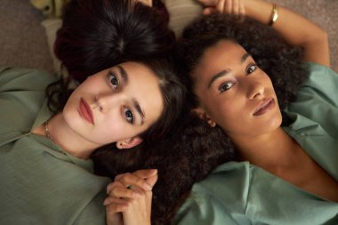 Yukarıdan bakıldığında, yerde uzanmış, el ele tutuşmuş ve kameraya bakan melez genç kız arkadaşları görülüyor.