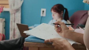 Sınıftan önce yatakhane odasının ahşap masasında matematik ödevi yapan, arka planda dizüstü bilgisayar kullanan bilinmeyen öğrencinin yakın görüntüsü.