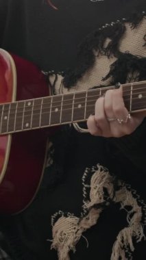 Yatakhanede akustik gitar çalan, yüz piercingli, yırtık kazaklı kadın zoomer görüntüsü.