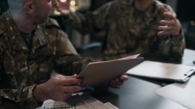 Kamuflaj üniformalı iki erkek komutanın askeri merkezde çalışırken belgeleri tartışmalarını yavaş çekimde kaldır.